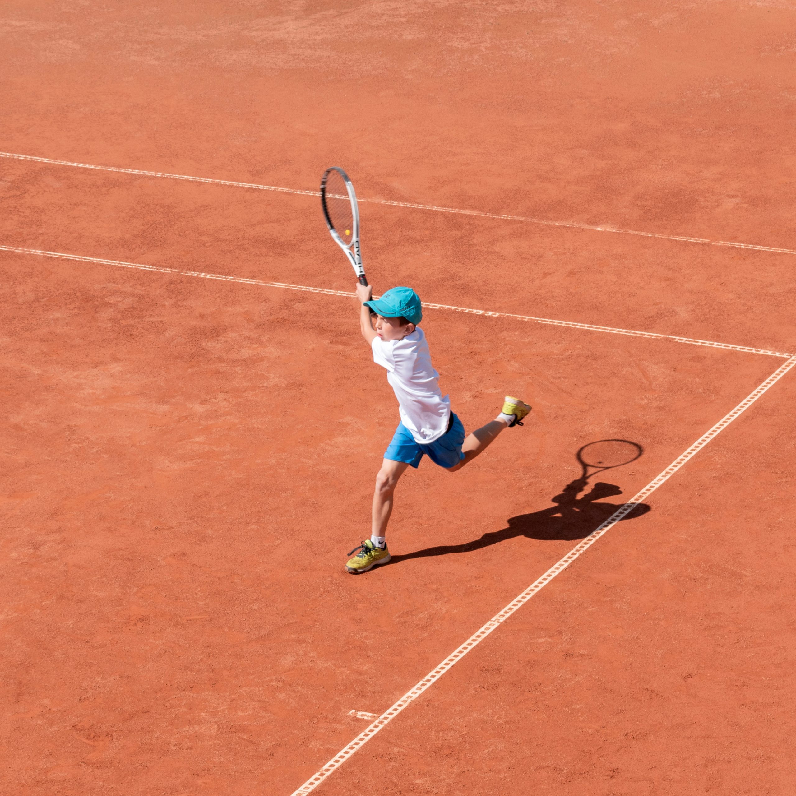 A boy plays tennis on a clay court. A little tennis player focused on the game and shot in flight after hitting the ball. Sports action frame. Active games. Square size.
