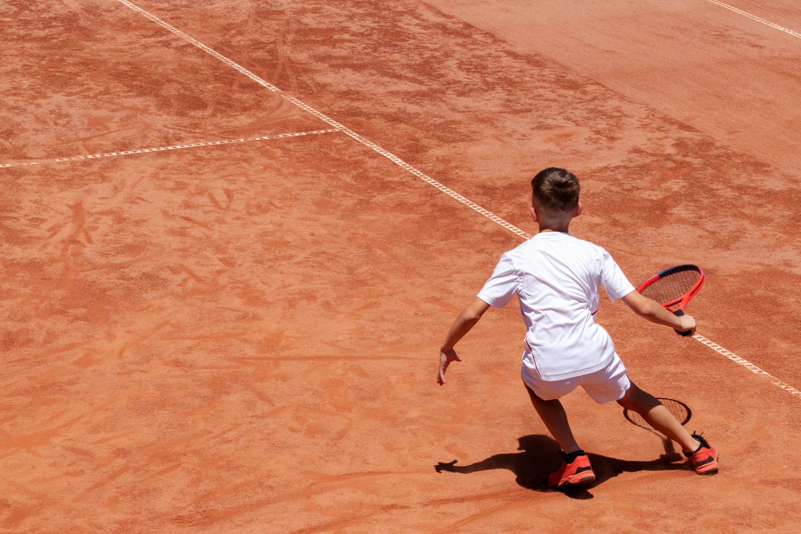 Young male tennis player with racket in action. Boy plays tennis on a clay tennis court. Child is concentrated and focused on the game. Kids tennis sport background with shadow. Motion. Copy space